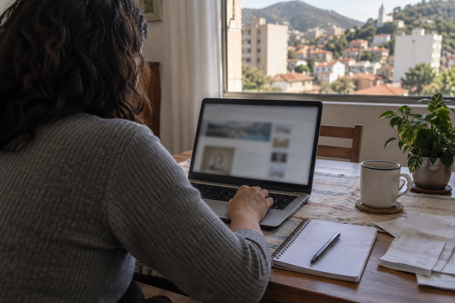 Pessoa de costas consulta um notebook com caderno e comprovantes sobre a mesa, em ambiente residencial de Minas Gerais.
